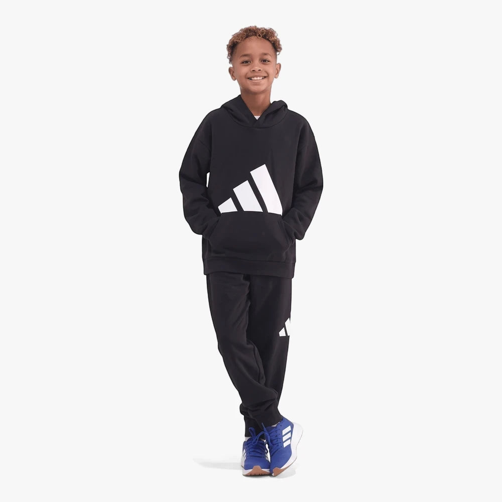 Young boy models a black Adidas BL FT Hoodie and sweatpants set, paired with blue sneakers, smiling with hands in pockets.