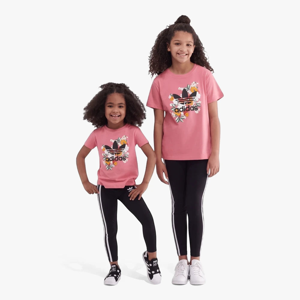 Two girls in pink Adidas Kids Floral Short Sleeve Tees with a colorful trefoil logo, black striped leggings, and sneakers.