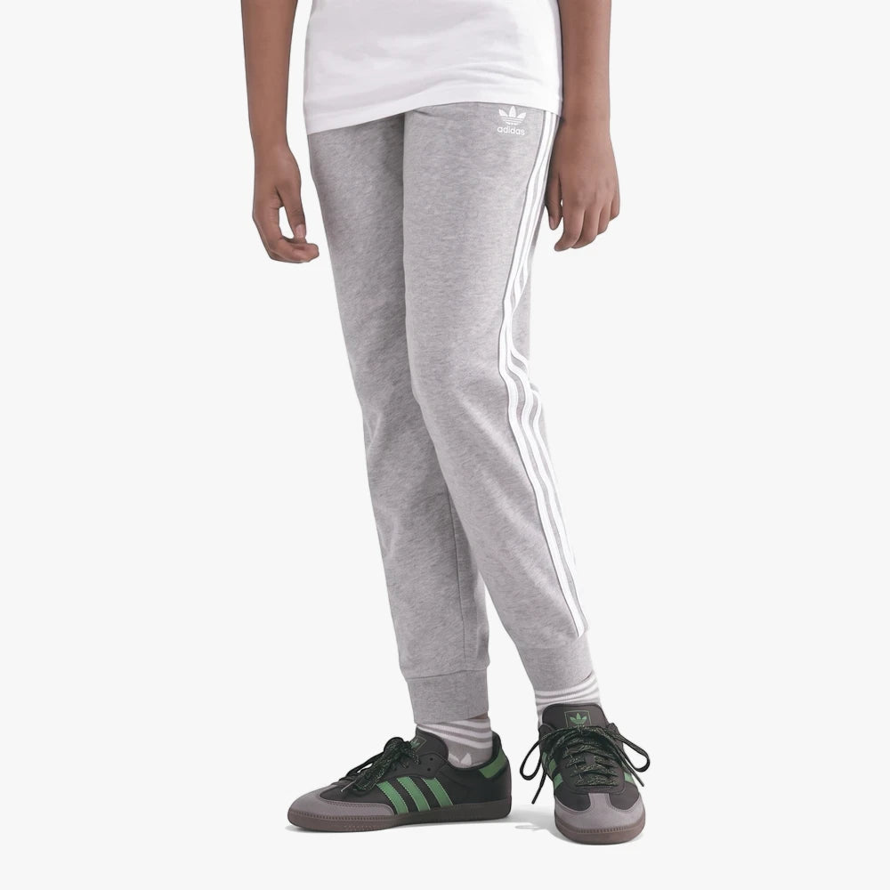 Adidas Youth Tref Pants in grey with signature three white stripes, worn by a youth model paired with dark sneakers.