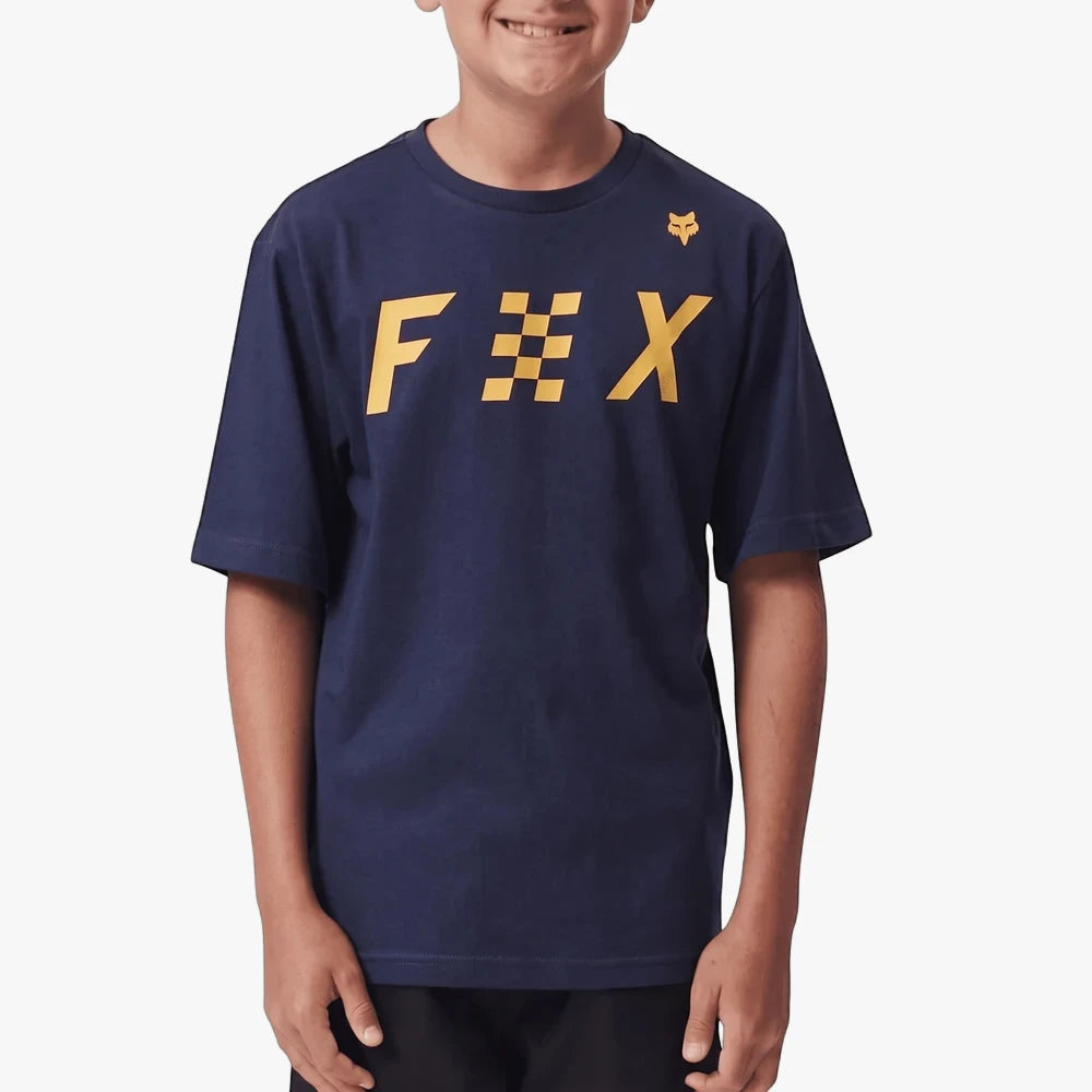 A boy wears a navy blue Fox Checkered Short Sleeve Tee. The shirt features a yellow Fox logo with a checkered E design.