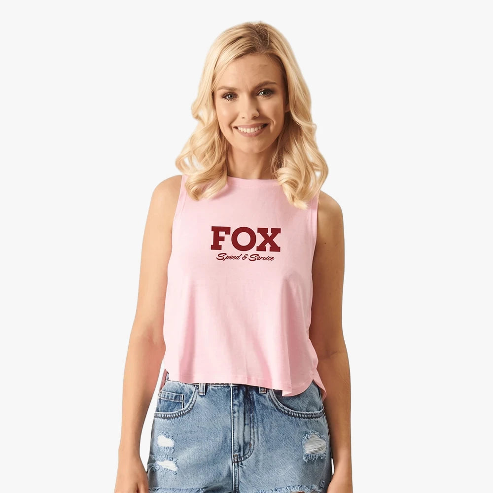 A smiling blonde woman wears a pink Fox Womens Speed Crop Tank Flamingo with a dark red logo, paired with distressed denim