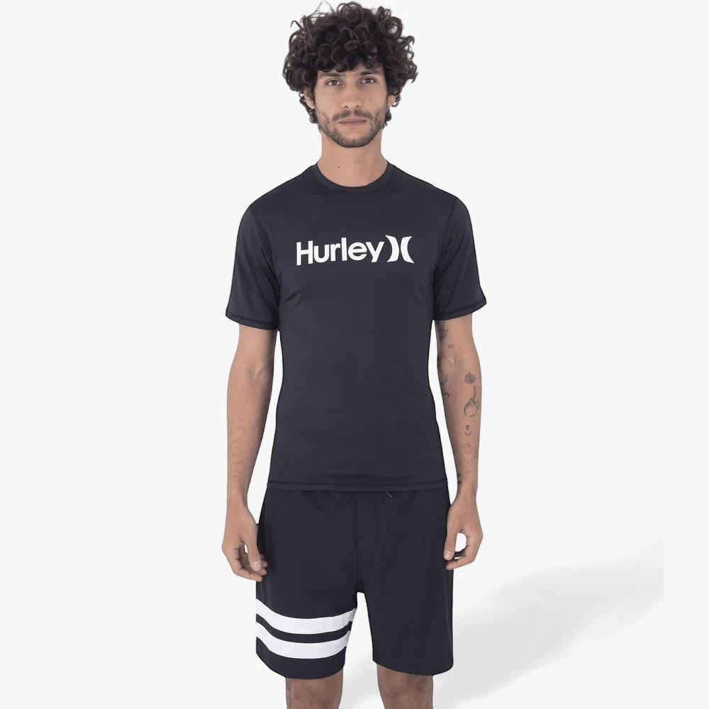 Male model wearing a black Hurley Oao Quickdry short sleeve rashguard and black shorts with white stripes, standing upright.