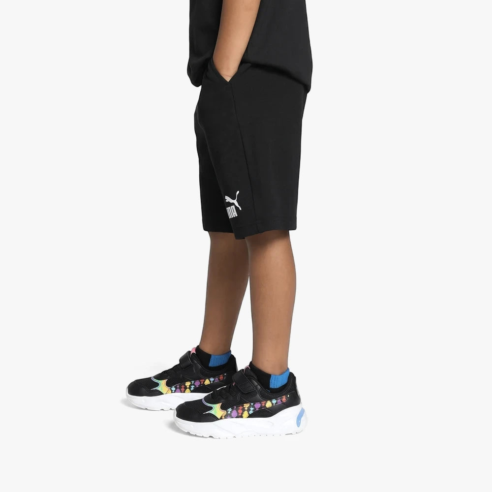 A boy wearing black Puma Essentials Jersey shorts, vibrant colorful sneakers, and blue socks, standing against a white