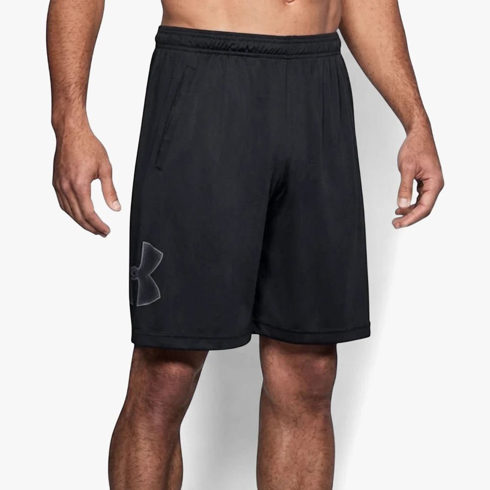 A man models black Under Armour Tech Graphic Shorts, featuring a gray logo on the left leg and an elastic waistband.