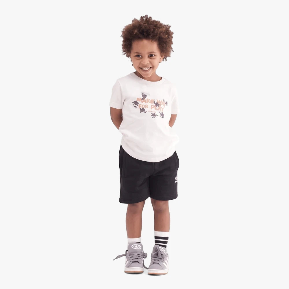 Young boy wearing Adidas Boys Logo Play 3 short sleeve tee in worn white beige, black shorts, and grey sneakers.