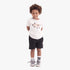 Young boy wearing Adidas Boys Logo Play 3 short sleeve tee in worn white beige, black shorts, and grey sneakers.