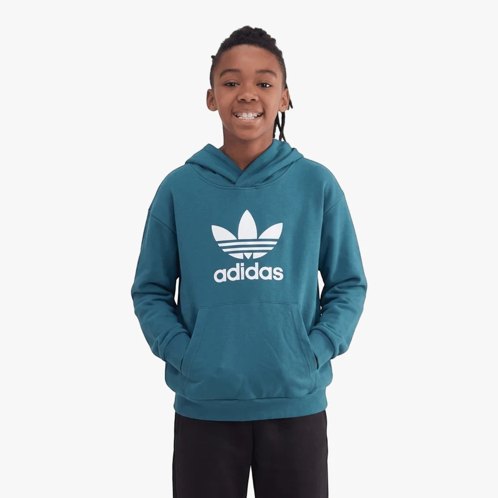 A smiling young boy with braided hair, wearing a teal Adidas Trefoil hoodie with a front kangaroo pocket and black pants.