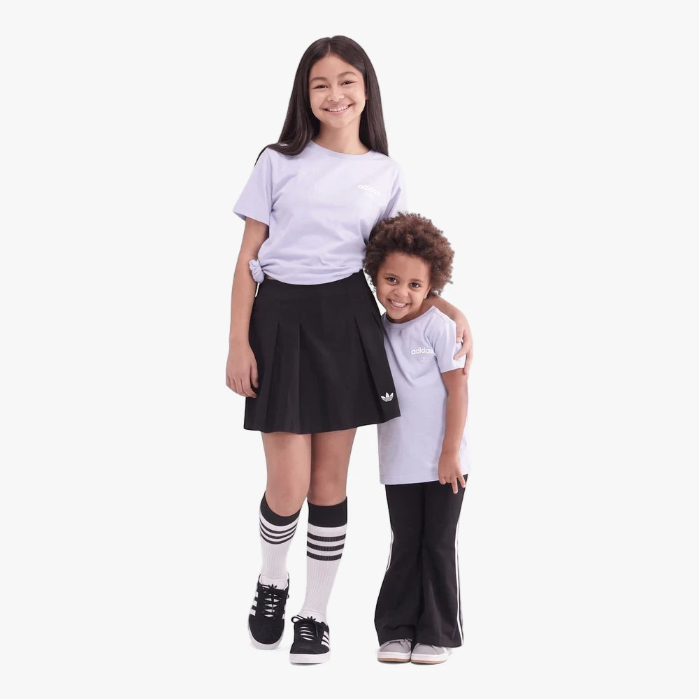 Two girls wearing matching Adidas Girls Varsity Short Sleeve Tee in violet, black skirts/pants, and sneakers, smiling in a