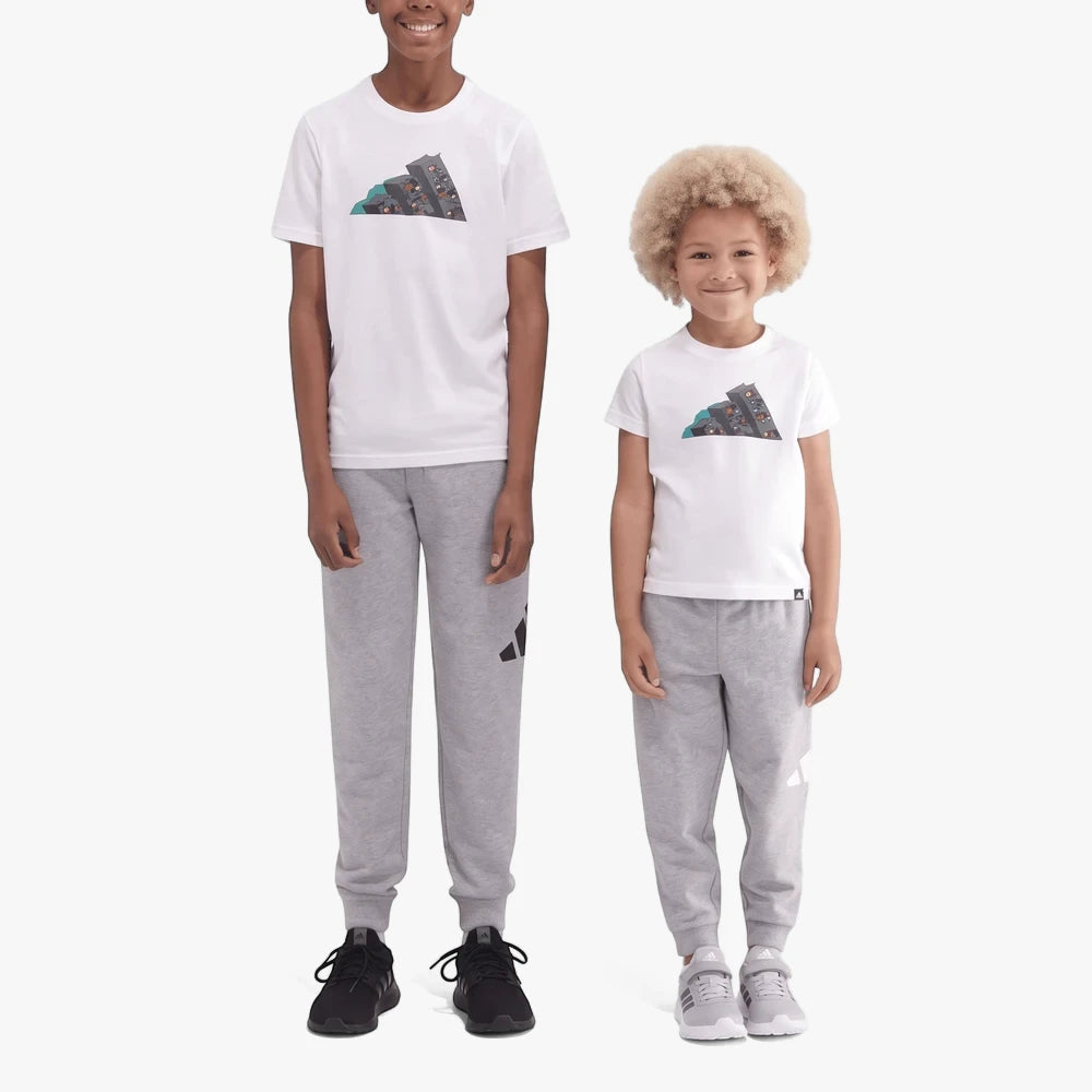Two kids wear white Adidas Kids Camo 2 Short Sleeve Tees with grey sweatpants and sneakers, standing against a white