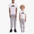 Two kids wear white Adidas Kids Camo 2 Short Sleeve Tees with grey sweatpants and sneakers, standing against a white