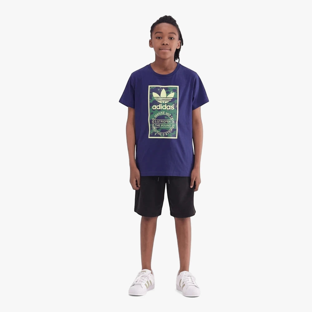 A young boy wearing a blue Adidas Kids Camo Tongue Short Sleeve Tee, black shorts, and white sneakers, standing against a