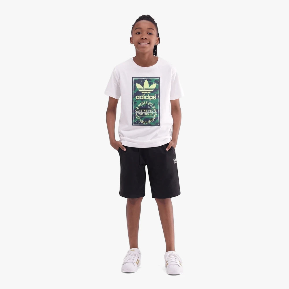Young boy wearing a white Adidas Kids Camo Tongue Short Sleeve Tee with black shorts and white sneakers, standing full body.