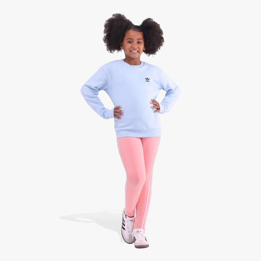 Young girl wearing a clear blue Adidas Kids Crew Ess sweater and pink three-stripe leggings, full-body studio shot.