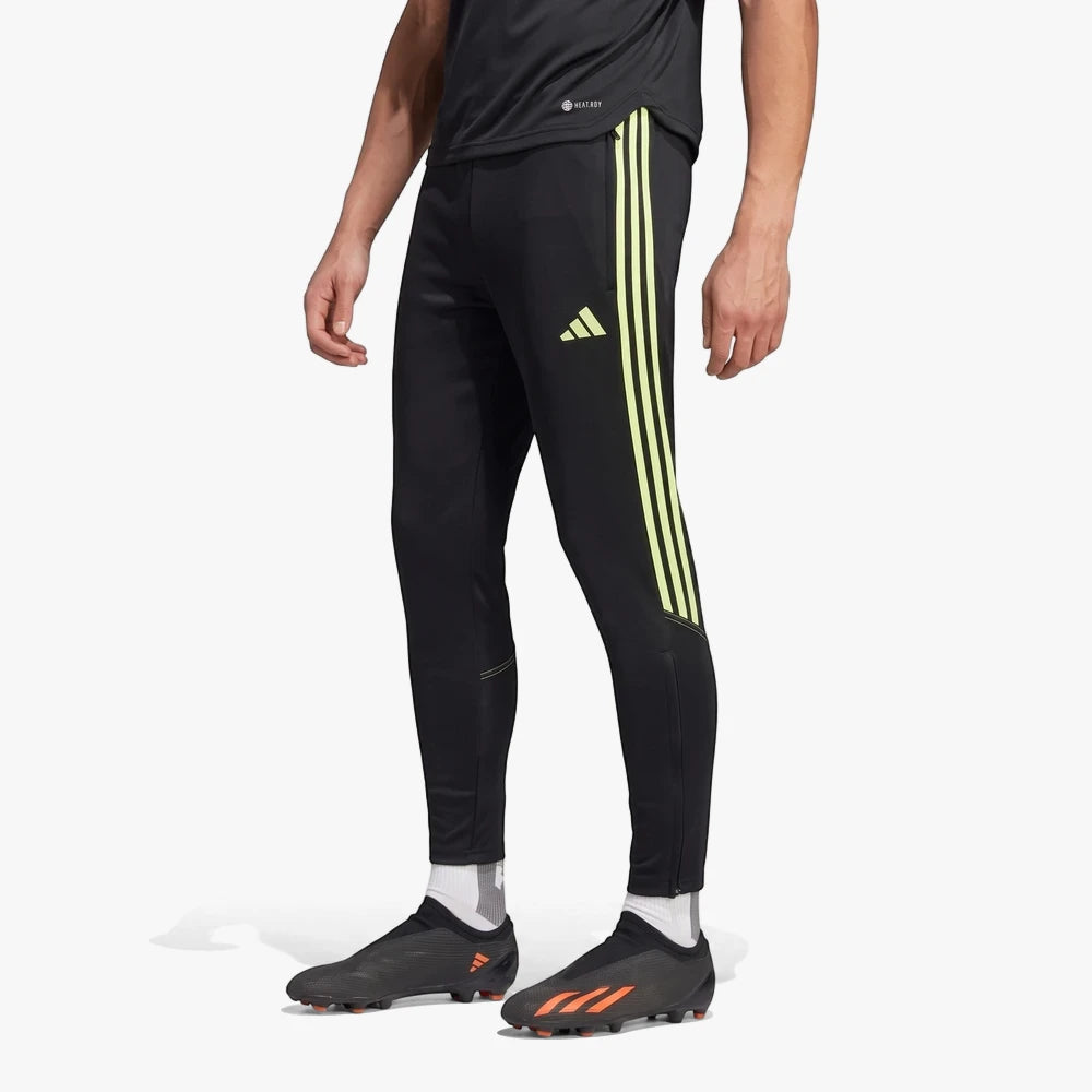 A man wears black Adidas Mens Tiro23 Trackpants with bright yellow-green stripes and black football boots on a light