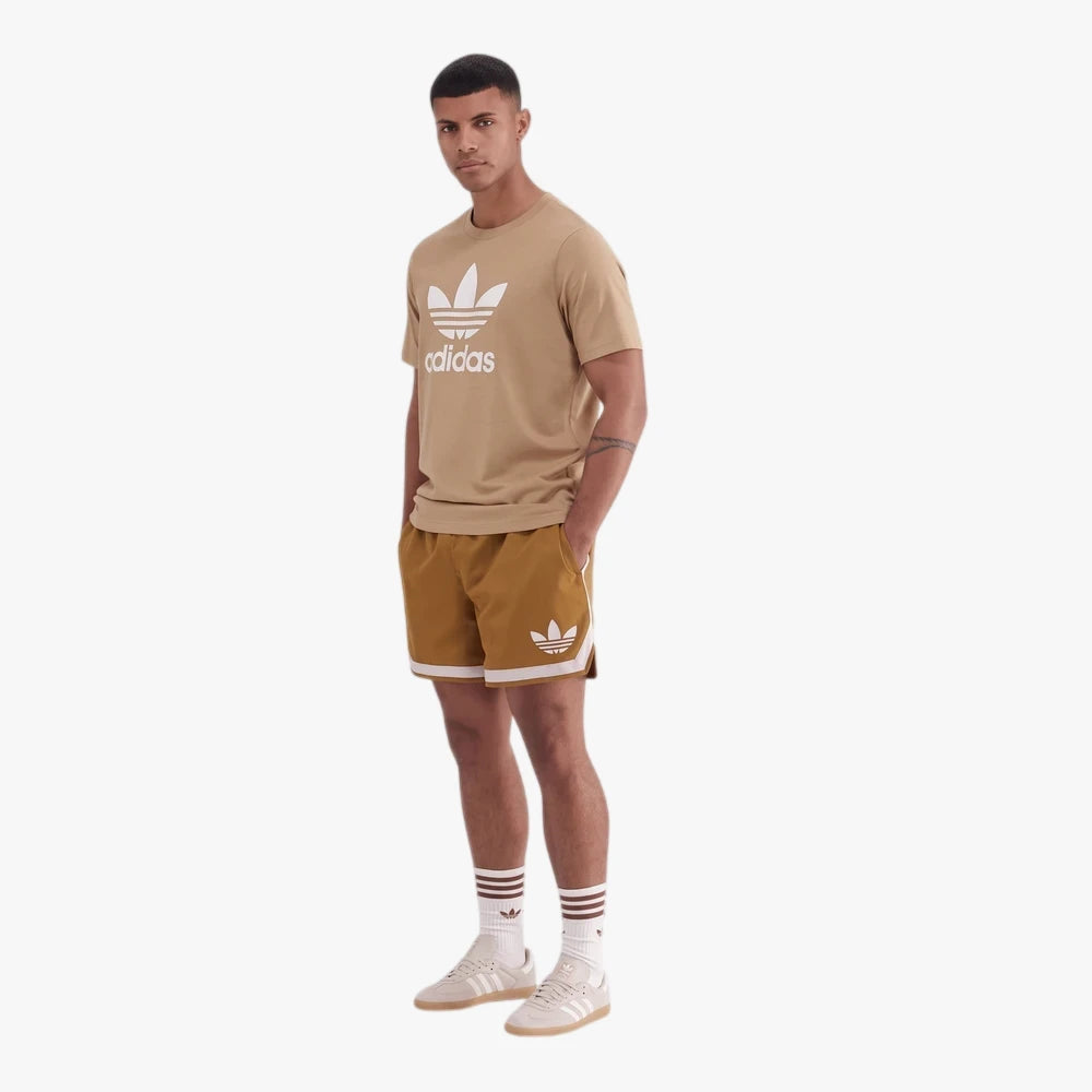 Male model wearing Adidas Mens Tref Short Sleeve Tee in cardboard color and matching shorts, socks, and sneakers, standing