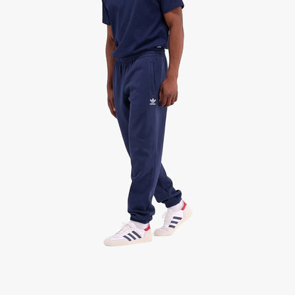 A man models dark blue Adidas Mens Trefoil Essentials French Terry Joggers with a white Trefoil logo, paired with white