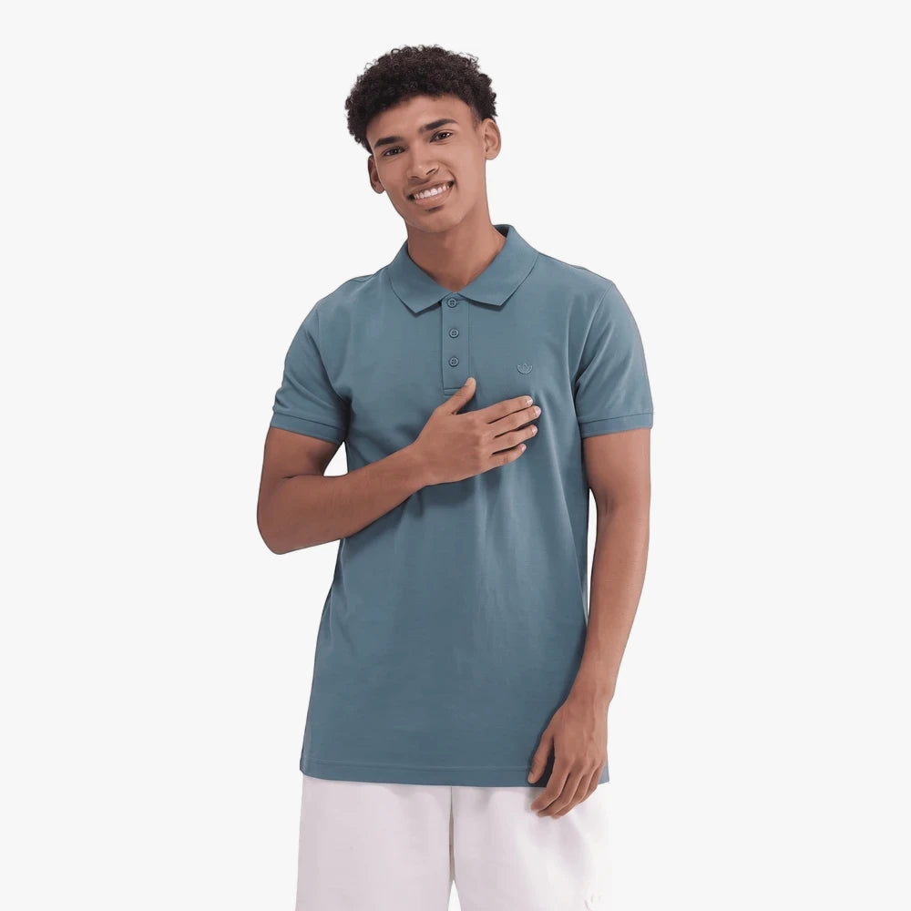 Adidas Mens Trefoil Essentials Polo Tee in a teal green shade, worn by a smiling man with curly hair and white shorts.