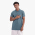 Adidas Mens Trefoil Essentials Polo Tee in a teal green shade, worn by a smiling man with curly hair and white shorts.