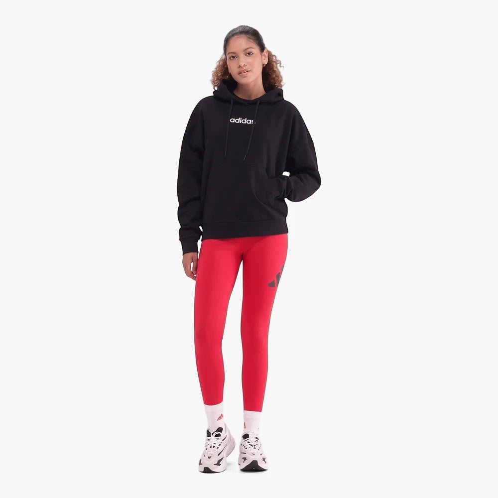 A woman models a black Adidas Lin Fl Hoodie paired with vibrant red leggings and sneakers, captured in a full-body studio