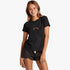 A young girl wears a black Billabong Pro short sleeve tee featuring a metallic logo and black scalloped shorts, smiling.