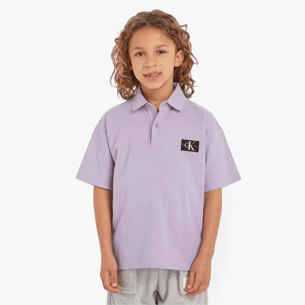 Young boy wearing a lavender Calvin Klein Kids Pique Badge Golfer polo shirt with the CK logo, paired with gray shorts.