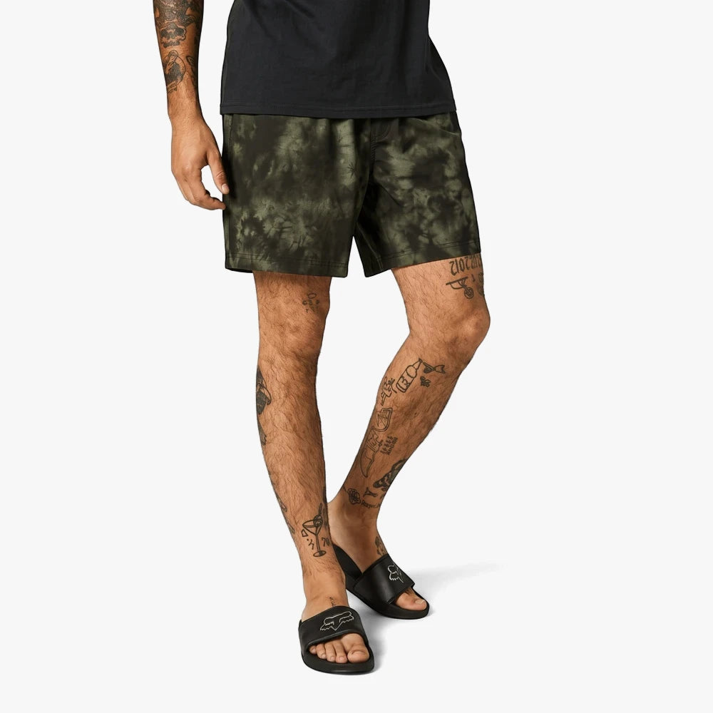 Fox Mens Essex Down N Dirty shorts in olive green tie-dye pattern, worn by a man with tattooed legs and black slide sandals.