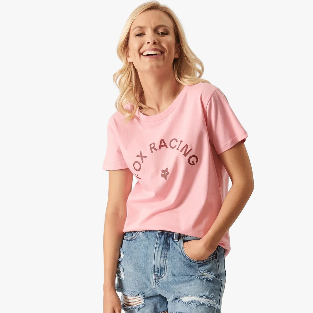 Smiling woman wearing a pink Fox Racing Arch short sleeve tee and distressed denim shorts, with hands in pockets.