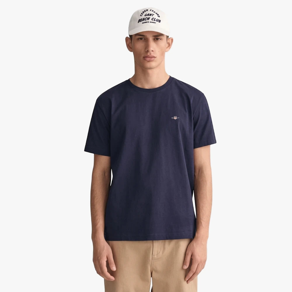 A male model wears a navy blue Gant Original Fit short sleeve tee, tan shorts, and a white baseball cap, standing against a