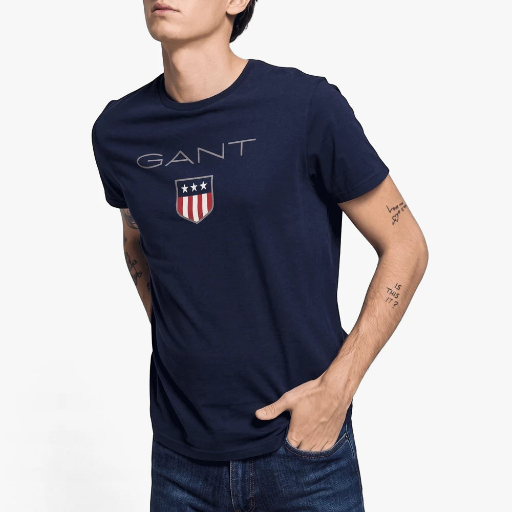 A male model wears a navy Gant Shield short sleeve t-shirt featuring the GANT logo and shield emblem, with hands in pockets.
