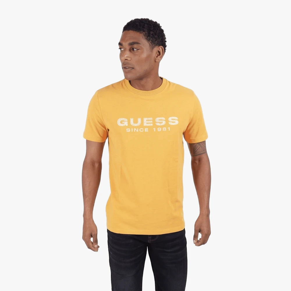 Male model wearing a yellow Guess short sleeve t-shirt with a white logo, paired with dark jeans, standing on a white