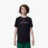 A young boy wears a black Nike Jdb Mj Wordmark Emb Crew short sleeve tee with white text and green shorts.