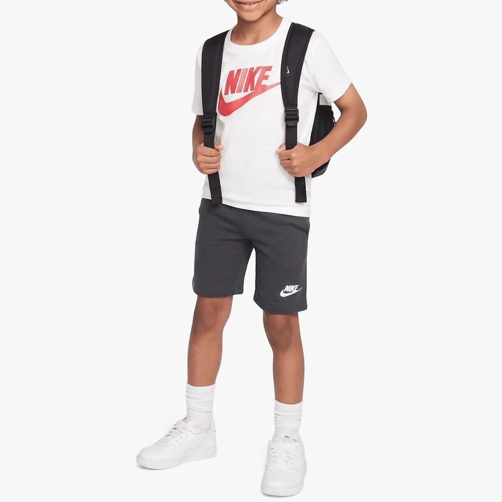 Young boy wearing a Nike Tape Shorts Set in anthracite, featuring a white Nike logo tee and a black backpack, standing on a
