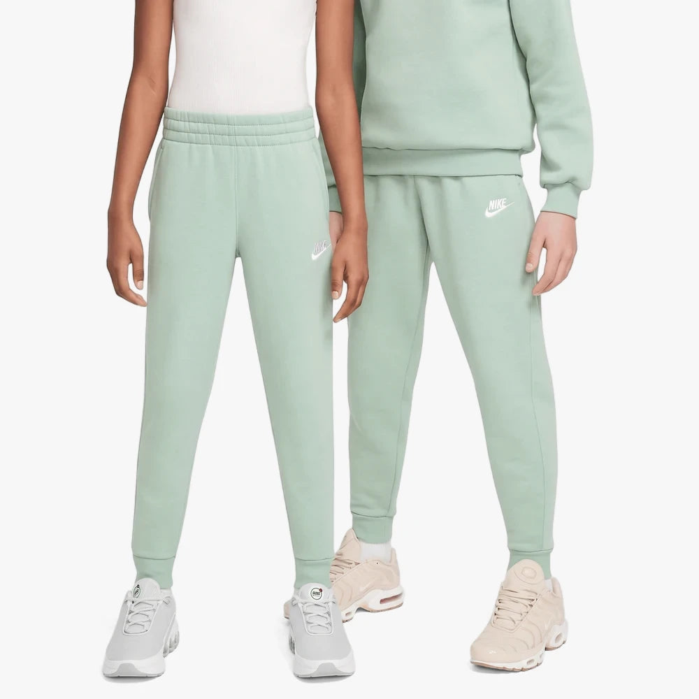 Two girls model comfortable Nike Girls Court Club Pants in sage green, featuring a white Nike logo and elastic cuffs. They