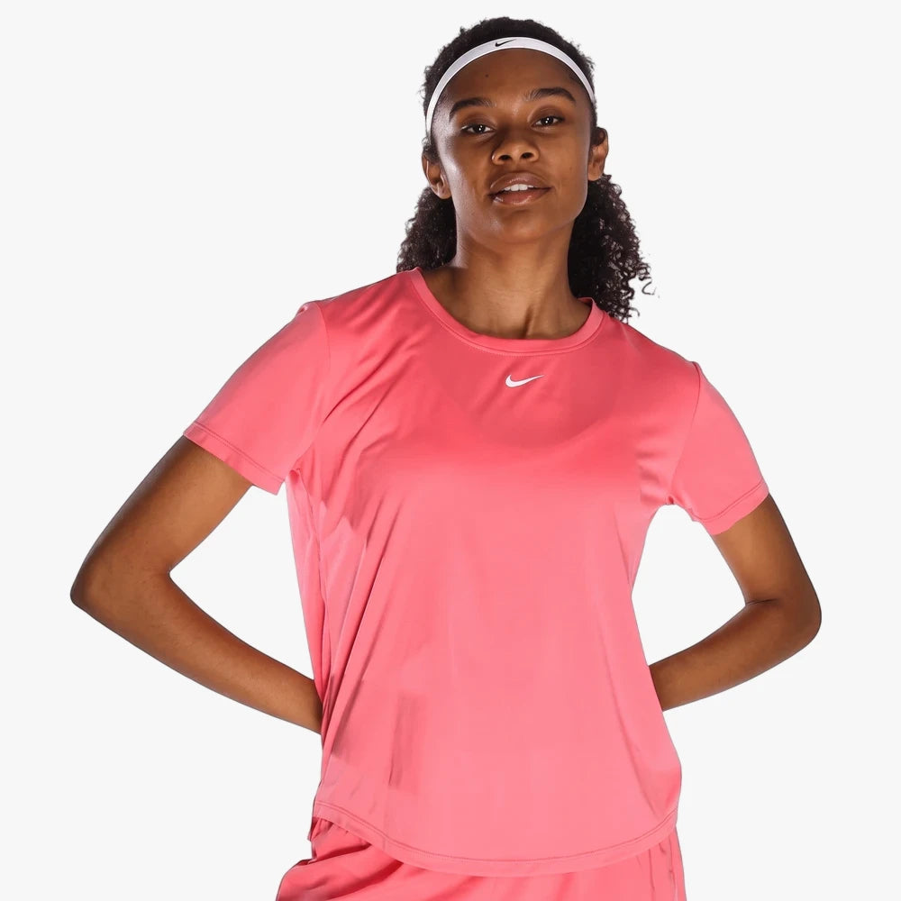 A woman wears a coral Nike Womens One Df Short Sleeve Tee and a white headband, standing with hands behind her back.
