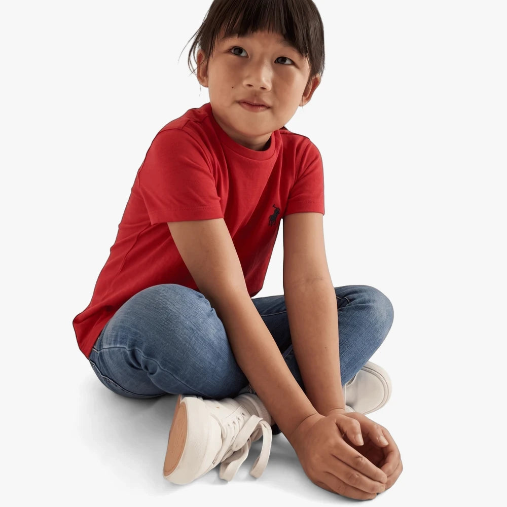 Young girl wearing a red short sleeve Polo Classic Tee, blue jeans, and white sneakers, sitting cross-legged and looking