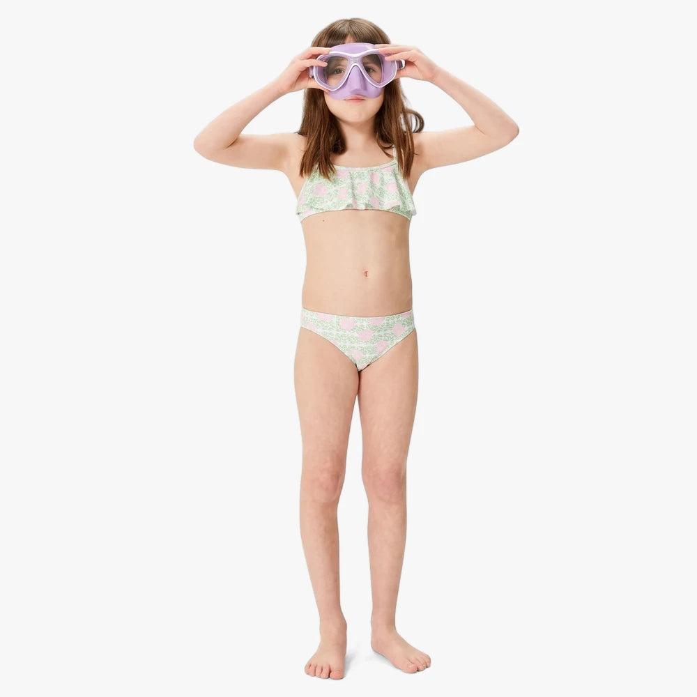 Young girl wearing a Roxy Girls Fairy Beach Flutter Set Sunshine Little Hibis light green and pink floral two-piece bikini,