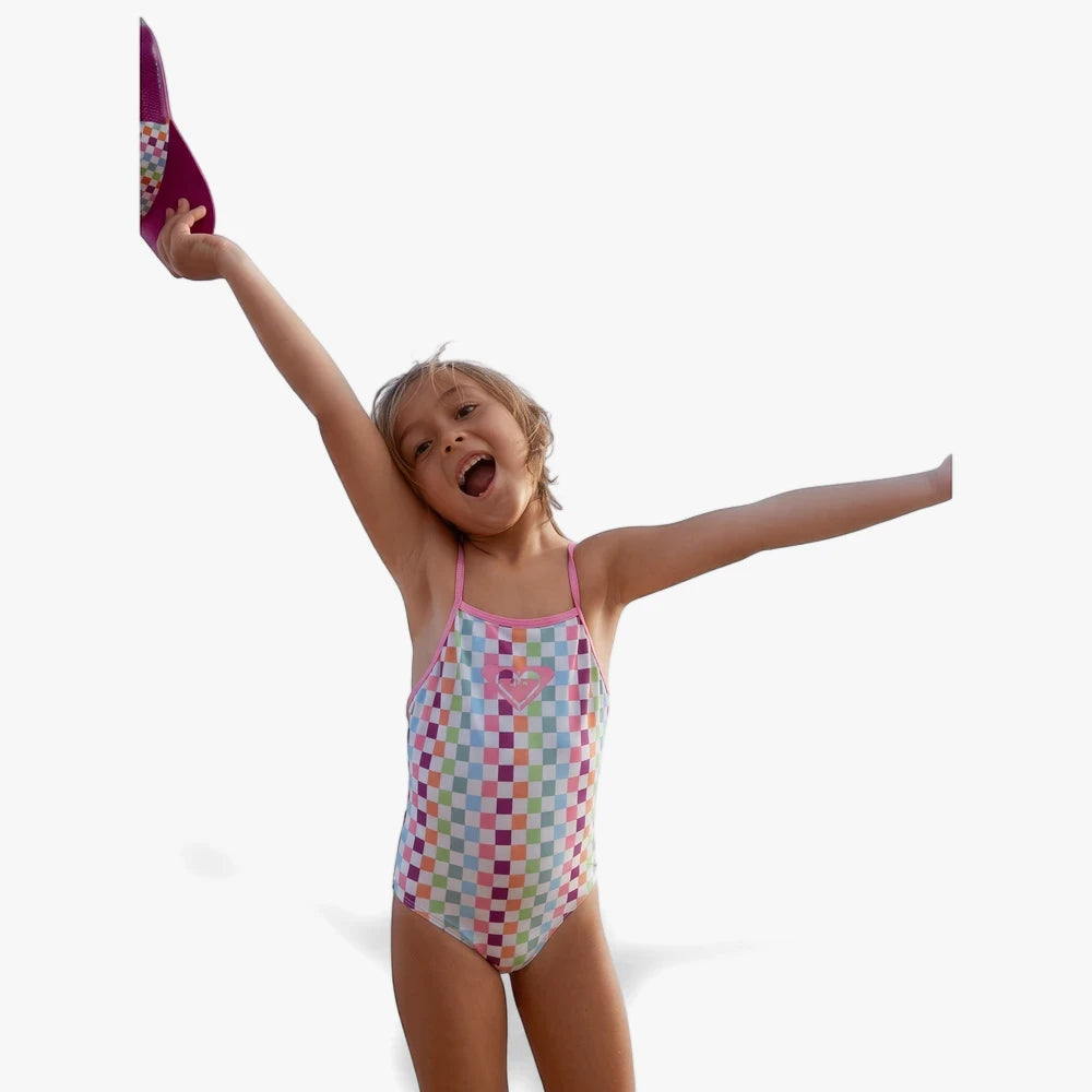 Young girl in a multi-color checkered Roxy one-piece swimsuit, raising her arms joyfully while holding a purple flip-flop.