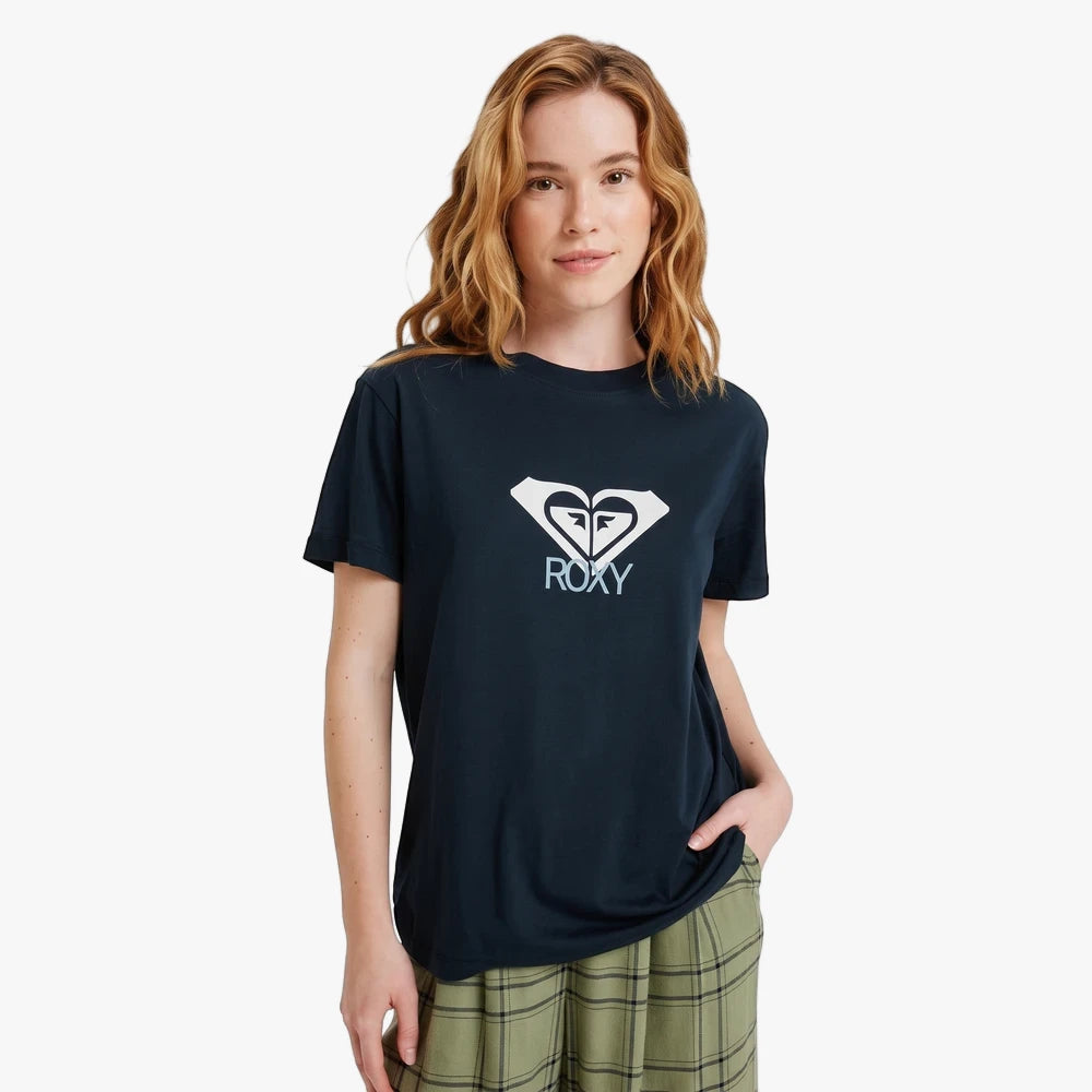 Young woman with wavy hair wearing a dark blue Roxy Ocean Road Art Tee featuring a white logo, paired with green plaid