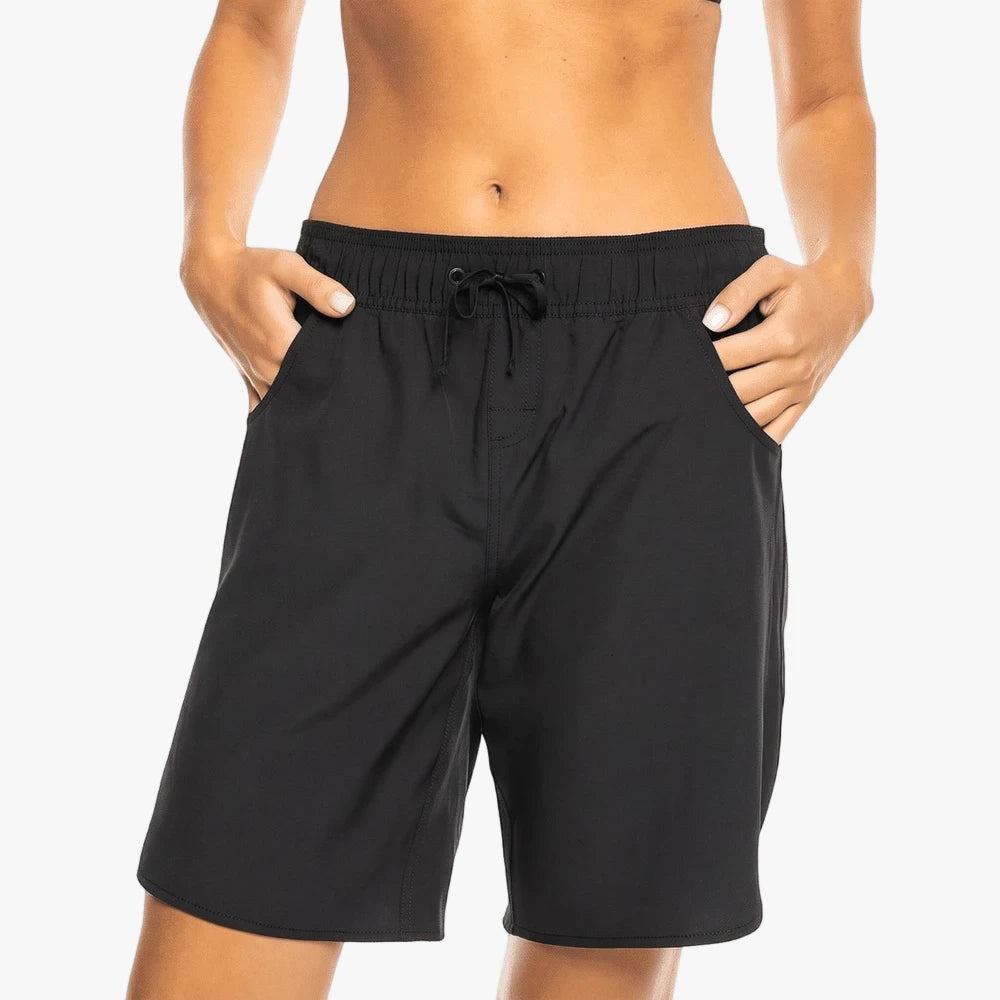 A close-up of a woman wearing black Roxy Wave 9" Shorts with a drawstring waist and hands in the pockets.