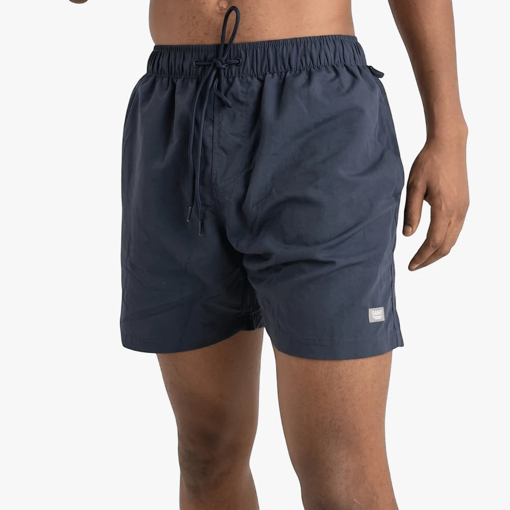 A man wears navy blue Saint And Summer Seth plain board shorts, showcasing the elastic drawstring waist and short leg length.