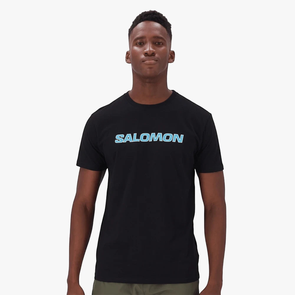A Black man wearing a black Salomon Talima short sleeve t-shirt with a light blue Salomon logo on the chest. He stands