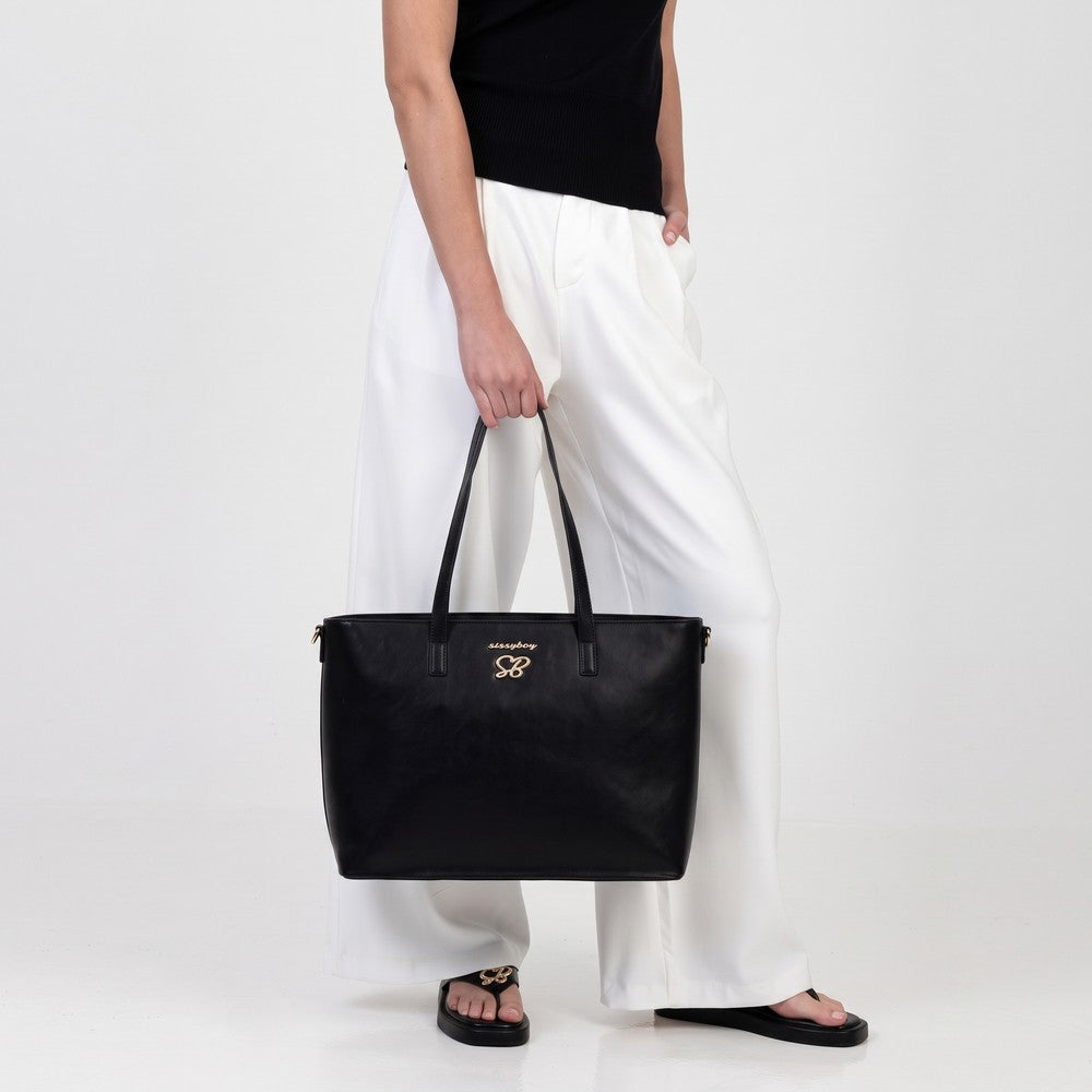 A woman holds a black Sissy Boy Collyns laptop bag with a gold logo. She wears a black top, white wide-leg pants, and black