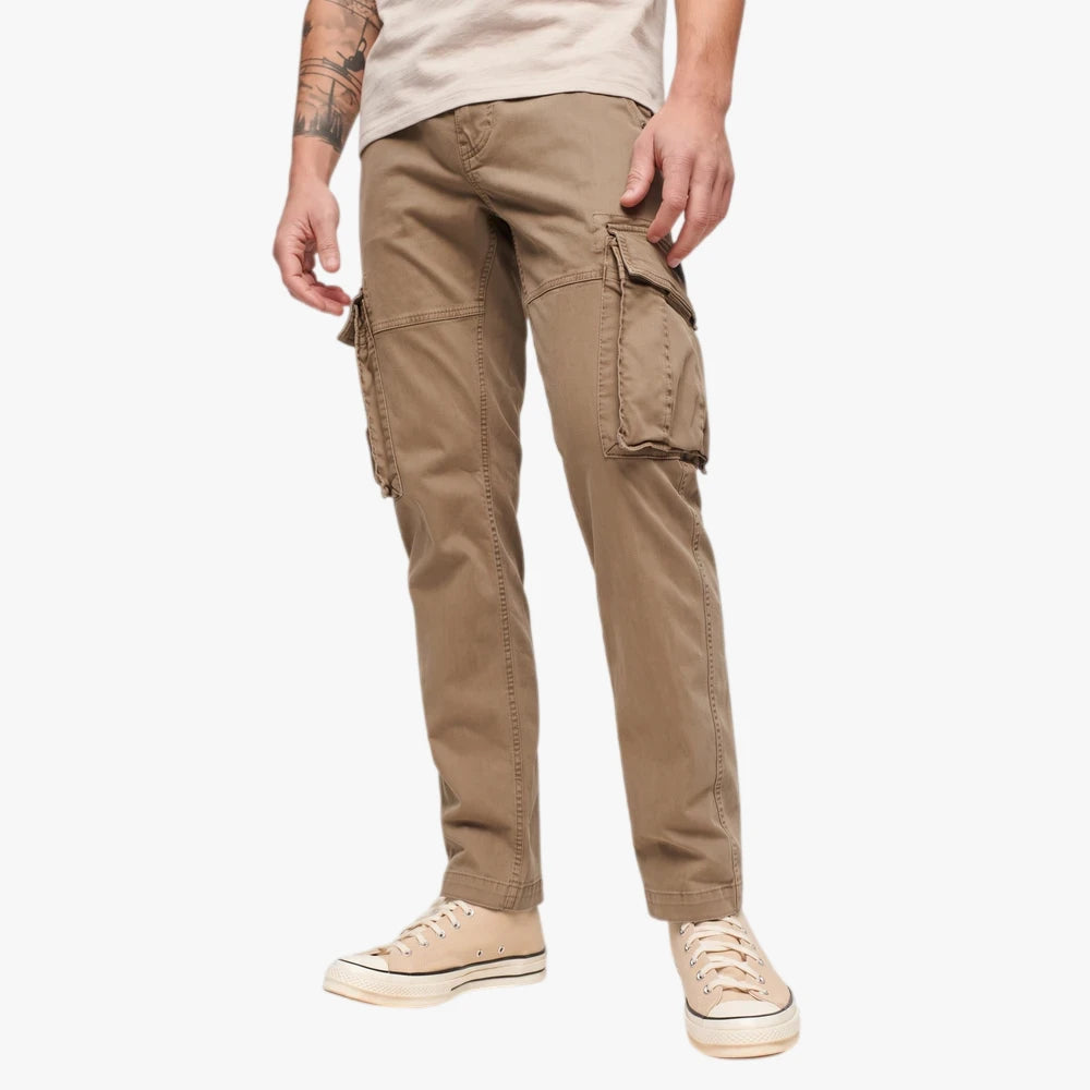 A person wears khaki Superdry Mens Baggy Cargo Pants with multiple flap pockets and beige canvas sneakers, standing in a