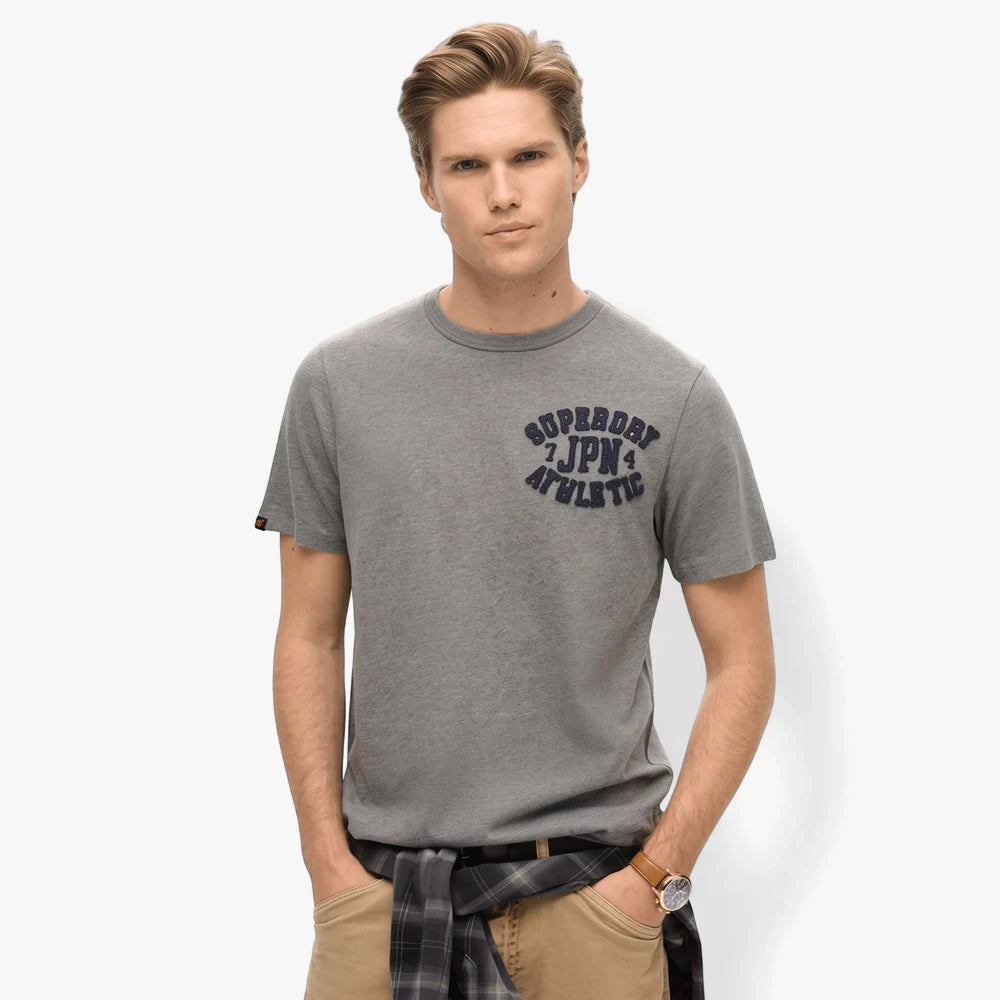 Male model wearing a grey Superdry Vintage Athletic Chest Tee, with a plaid shirt tied around his waist and khaki pants,