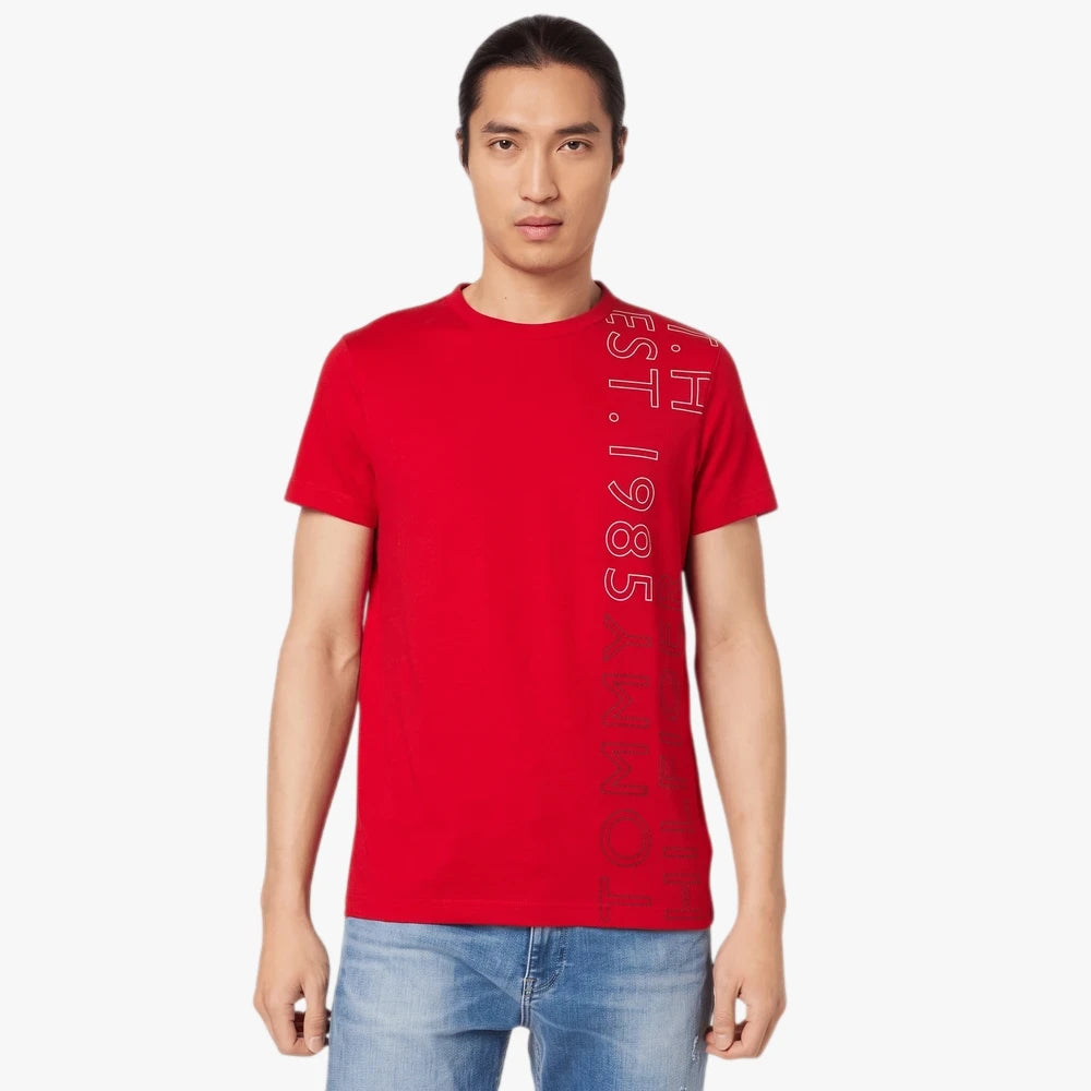 Male model wearing a red Tommy Hilfiger Off Placement Tee with vertical text and blue jeans, standing in a studio.