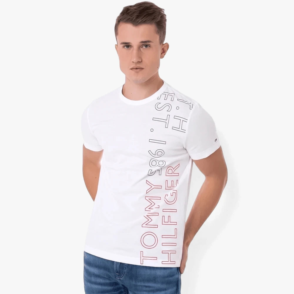 A male model wearing a white Tommy Hilfiger Off Placement tee with a vertical brand graphic and blue jeans.
